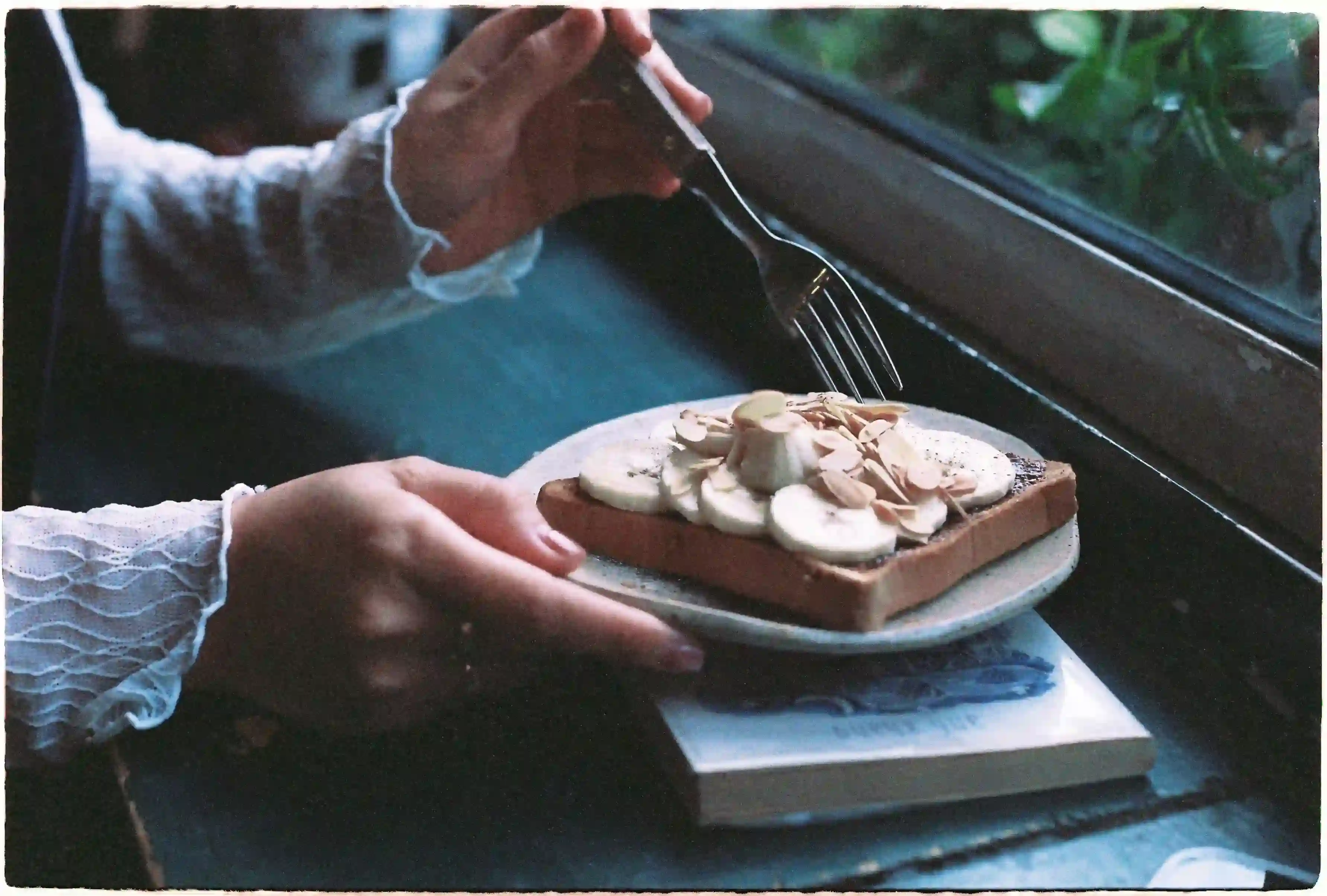 Person holding a fork over a plate with a slice of toast topped with banana slices, almonds, and a dollop of cream, placed on a book at a table by a window, inspiring healthy eating habits for how to start intermittent fasting