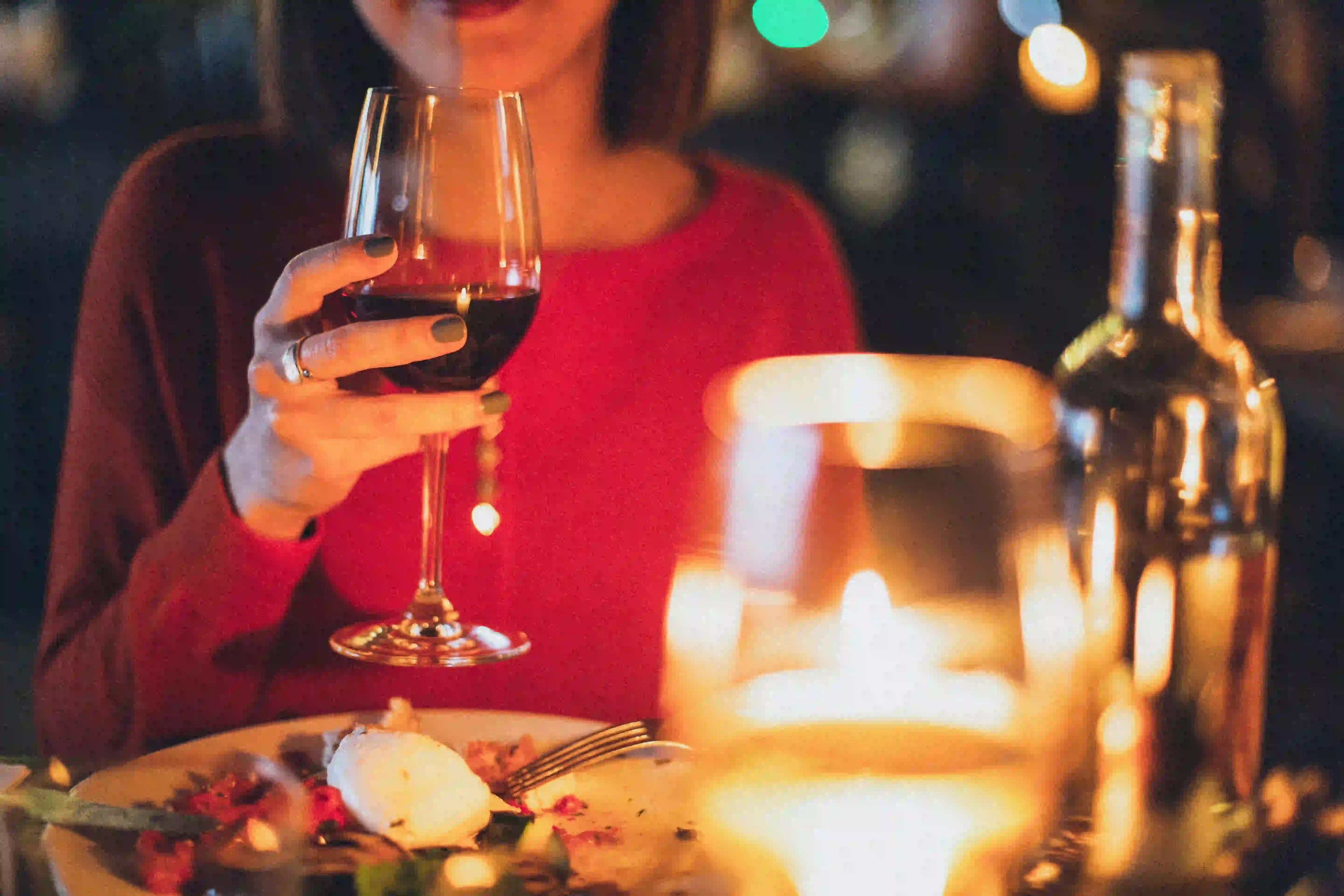 Person in a red sweater holding a glass of red wine at a cozy dinner table with mozzarella and vegetables, suitable for intermittent fasting plans like skipping breakfast or dinner