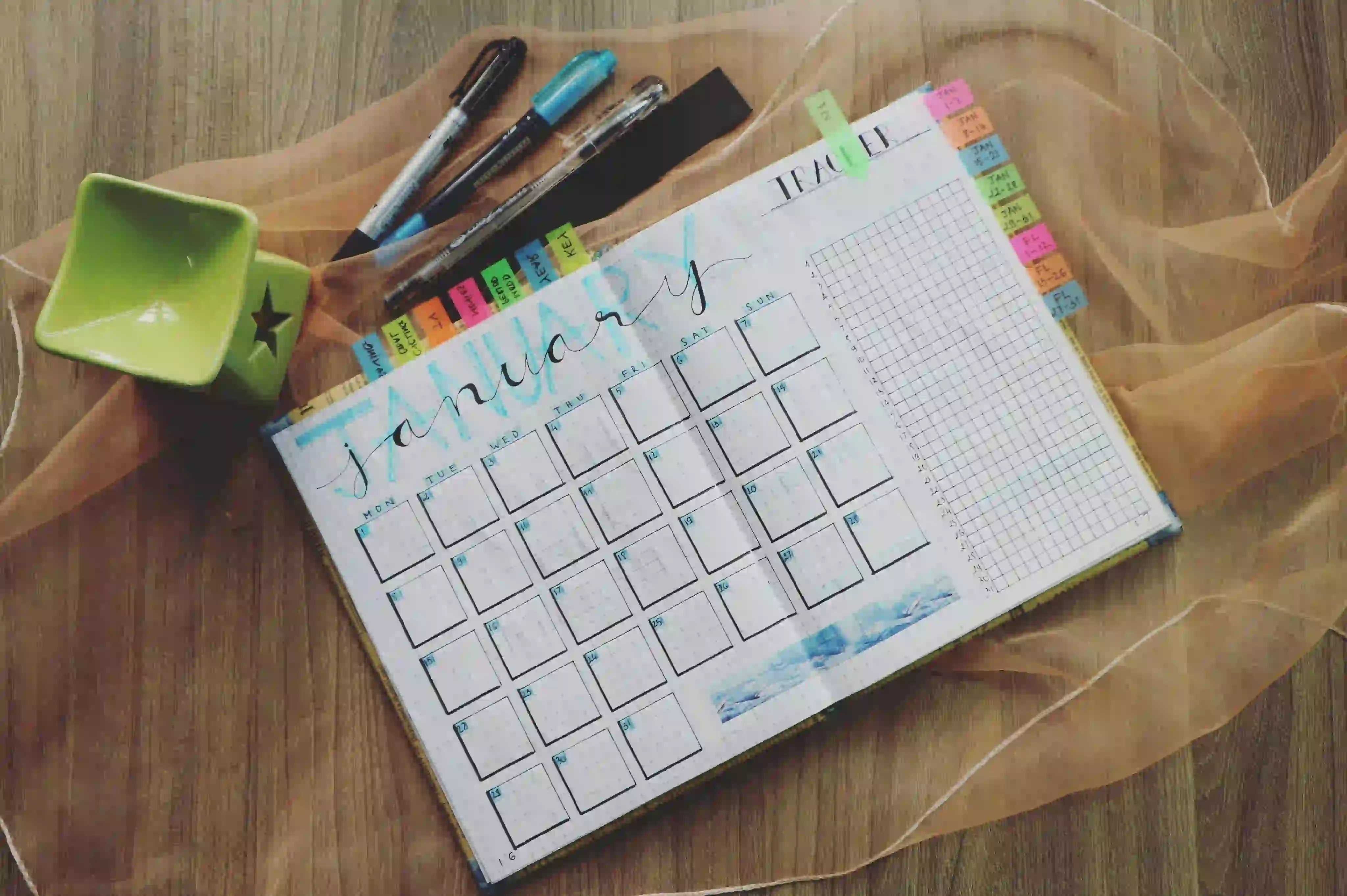 Open January planner on a wooden table with a calendar, pens, green candle holder, and sticky notes, ideal for scheduling intermittent fasting plans like skipping breakfast or dinner