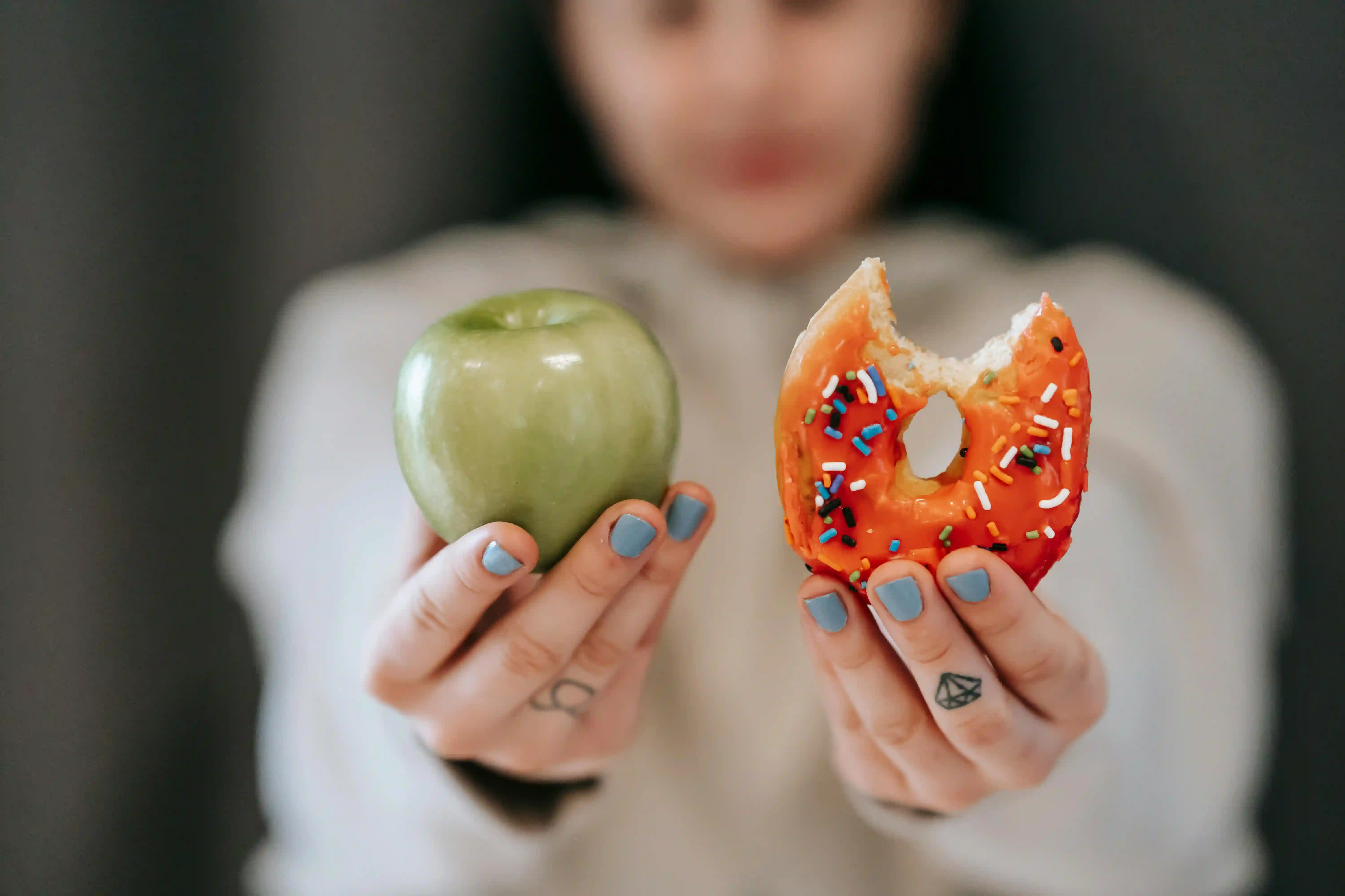 A person follows intermittent fasting rules, holding a green apple in one hand and a bitten, sprinkled donut in the other, both hands extended toward the camera