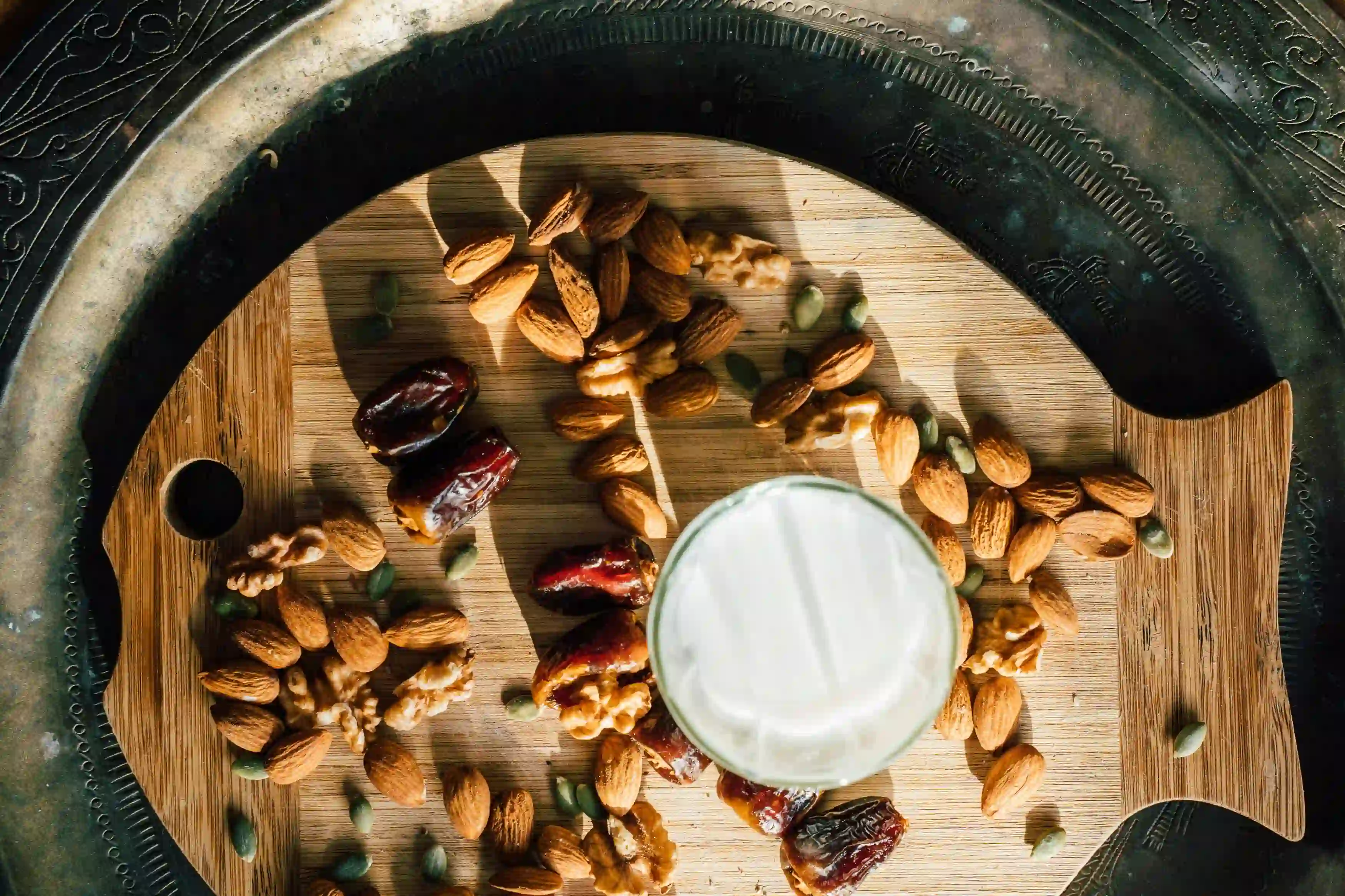 A wooden tray shaped like a fish holding a variety of nuts including almonds, walnuts, and pumpkin seeds, along with dried dates, and a glass of white liquid, possibly milk or yogurt, promoting healthy intermittent fasting rules